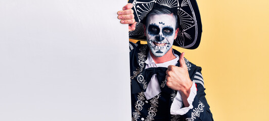 Young man wearing mexican day of the dead costume holding blank empty banner smiling happy and positive, thumb up doing excellent and approval sign