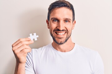 Young handsome man holding piece of puzzle standing over isolated white background looking positive and happy standing and smiling with a confident smile showing teeth