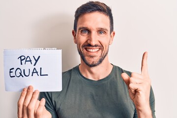 Young handsome man asking for equality economy holding paper with pay equal message smiling happy pointing with hand and finger to the side