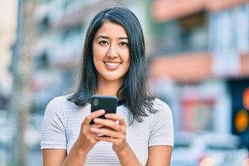 Fototapeta premium Young hispanic woman smiling happy using smartphone at the city.
