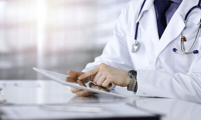 Unknown male doctor sitting and working with tablet computer iin a darkened clinic, glare of light...