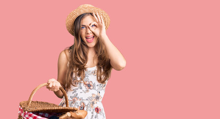 Beautiful caucasian young woman wearing summer hat and holding picnic wicker basket with bread smiling happy doing ok sign with hand on eye looking through fingers