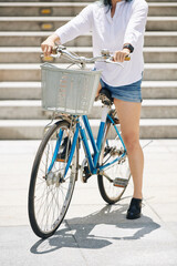 Cropped image of young woman in denim shorts sitting on bicycle with big front basket