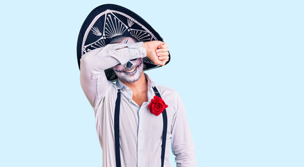 Man wearing day of the dead costume over background covering eyes with arm smiling cheerful and funny. blind concept.