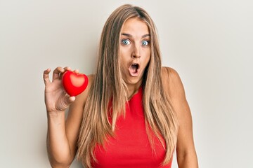 Young blonde woman holding heart scared and amazed with open mouth for surprise, disbelief face