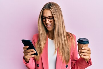 Young blonde woman using smartphone and drinking a cup of coffee winking looking at the camera with...