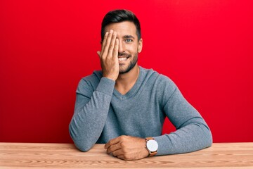 Fototapeta premium Handsome hispanic man wearing casual style sitting on the table covering one eye with hand, confident smile on face and surprise emotion.