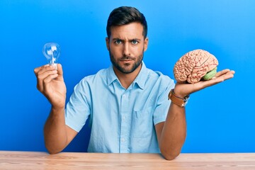 Handsome hispanic man holding lightbulb and brain for inspiration and idea skeptic and nervous,...