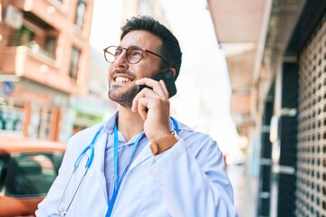 Obraz premium Young handsome hispanic doctor wearing stethoscope smiling happy Standing with smile on face talking on the smartphone at town street.