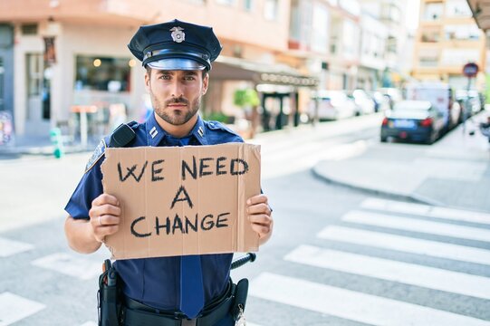 Young handsome hispanic policeman wearing police uniform with serious expression holding we need a change banner at town street