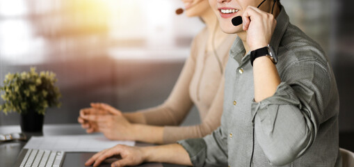 Fototapeta premium Two smiling people in headsets are talking to the clients, while sitting at the desk in sunny modern office