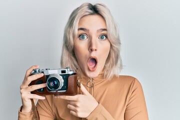 Young blonde girl holding vintage camera afraid and shocked with surprise and amazed expression, fear and excited face.