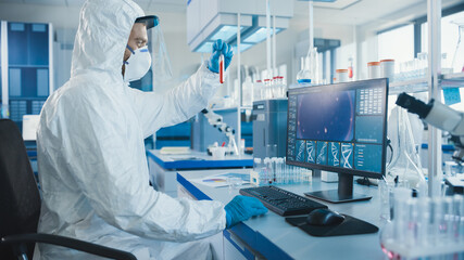 Microbiology Laboratory: Scientist Wearing Sterile Coverall, Face Shield and Mask Works on Computer Developing Vaccine, Drugs, Antibiotics Holds Test Tube with Blood Sample.Screen Shows DNA 
