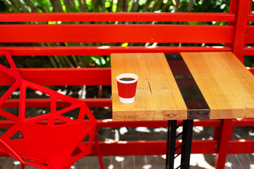 A wooden table on which there is a red plastic cup with coffee. Summer beautiful terrace in greenery. Cozy place.