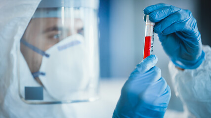 Medical Research Scientist Wearing Coverall, Surgical Gloves, Face Mask and Shield Holds Test Tube with Blood Sample and Label Reading Covid-19. Microbiology Laboratory Drug and Vaccine Development