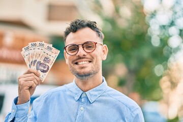 Young hispanic businessman smiling happy holding mexican 500 pesos banknotes at the city. - obrazy, fototapety, plakaty