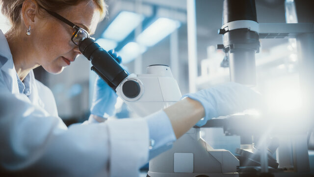 Bright Medical Science Laboratory: Beautiful Microbiologist Looks Under Microscope Does Analysis of Test Sample. Brilliant Scientist, Doctor, working with High-Tech Equipment. Close-up with Sun Flare.