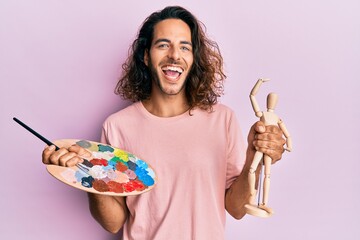 Young handsome man with long hair holding painter palette and art manikin celebrating crazy and amazed for success with open eyes screaming excited.
