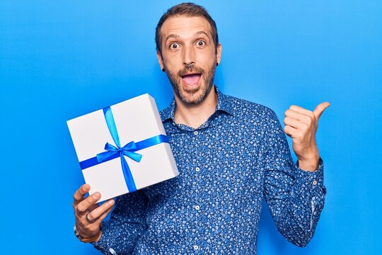 Young handsome man holding gift pointing thumb up to the side smiling happy with open mouth