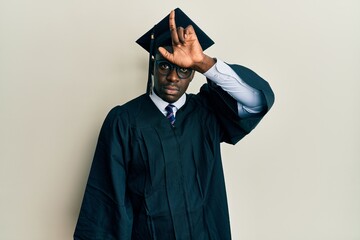 Handsome black man wearing graduation cap and ceremony robe making fun of people with fingers on forehead doing loser gesture mocking and insulting.