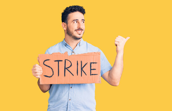 Young hispanic man holding strike banner cardboard pointing thumb up to the side smiling happy with open mouth