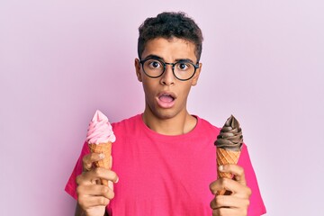 Young handsome african american man holding ice cream cones in shock face, looking skeptical and sarcastic, surprised with open mouth