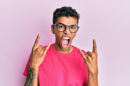 Young handsome african american man wearing glasses over pink background shouting with crazy expression doing rock symbol with hands up. music star. heavy concept.