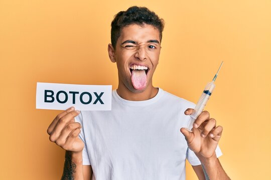 Young Handsome African American Man Holding Botox Banner And Needle Sticking Tongue Out Happy With Funny Expression.