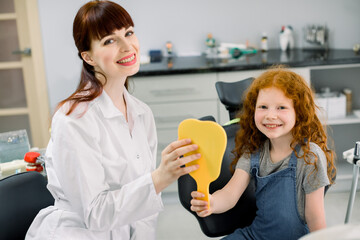 Little red haired girl sitting on dentist chair looking at camera and smiling while keeping yellow...