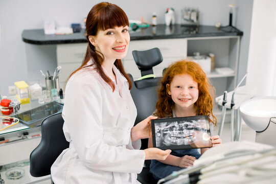Little smiling girl with red curly hair sitting on chair and looking at camera, while holding x-ray scan image of her teeth on digital tablet together with her cheerful female dentist at dental clinic
