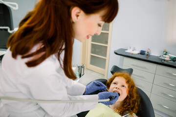 Top angle view of cute red haired girl lying on dentist chair with open mouth while young female doctor curing her teeth, using dental drill. Caries prevention and treatment. Child at dental clinic