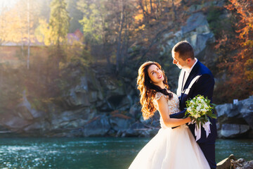 profile of newlyweds on the background of rocks and river. beautiful bride in white dress holds wedding bouquet and groom hugs her by the waist.