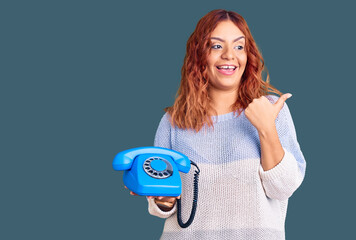 Young latin woman holding vintage telephone pointing thumb up to the side smiling happy with open mouth