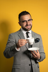 Cheerful young handsome bearded man in eye glasses in formal wear holding a cup of coffee isolated on yellow background. 