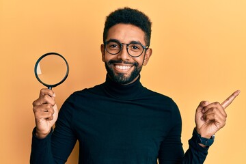 Handsome hispanic man with beard holding magnifying glass smiling happy pointing with hand and...