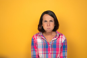 Fototapeta premium Making funny faces pretty looking girl looking to camera. Woman in a plaid shirt on yellow background. Human emotions, facial expression concept.