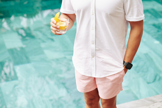 Cropped image of man in shirt and pink shorts standing by swimming pool and drinking gin and tonic