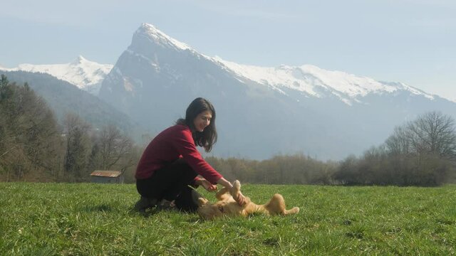 A gorgeous young woman plays with her Golden Retriever puppy in a field with a majestic mountain peak in the background, slow motion.