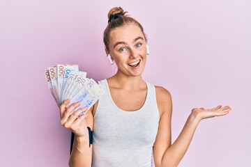 Beautiful blonde sport woman holding 20 swedish krona banknotes celebrating achievement with happy smile and winner expression with raised hand