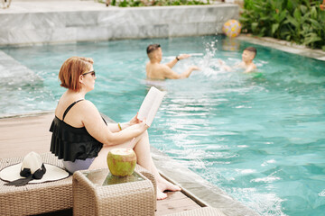 Woman sitting on chaise-lounge and reading book when her husband and son playing with inflatable ball in swimming pool