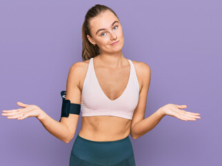 Fototapeta na wymiar Beautiful young blonde woman wearing sportswear and arm band clueless and confused expression with arms and hands raised. doubt concept.