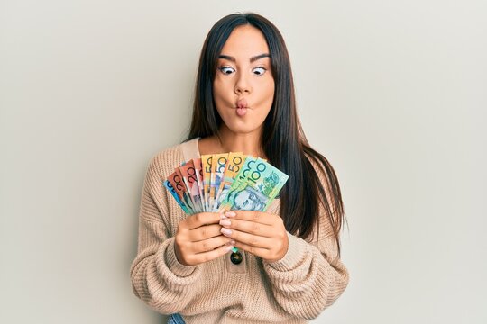 Young beautiful hispanic girl holding australian dollars making fish face with mouth and squinting eyes, crazy and comical.