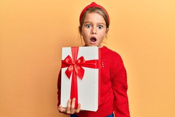Little beautiful girl holding gift scared and amazed with open mouth for surprise, disbelief face
