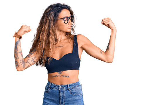 Young hispanic woman with tattoo wearing casual clothes and glasses showing arms muscles smiling proud. fitness concept.