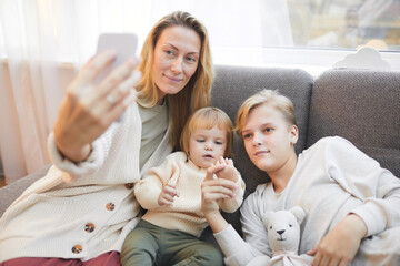 Portrait of modern mature mother taking selfie photo with two kids while relaxing together on sofa at home, copy space