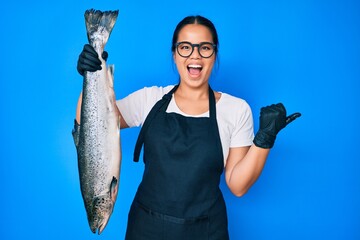 Young beautiful asian girl fishmonger selling fresh raw salmon pointing thumb up to the side...