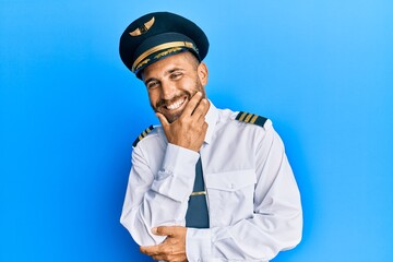 Handsome man with beard wearing airplane pilot uniform looking confident at the camera smiling with crossed arms and hand raised on chin. thinking positive.