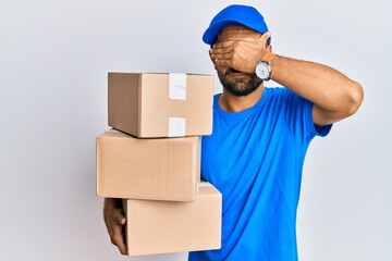 Handsome man with beard wearing courier uniform holding delivery packages covering eyes with hand,...