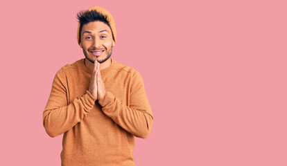 Fototapeta premium Handsome latin american young man wearing winter sweater and wool hat praying with hands together asking for forgiveness smiling confident.