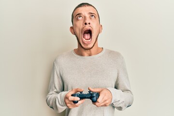 Hispanic young man playing video game holding controller angry and mad screaming frustrated and...